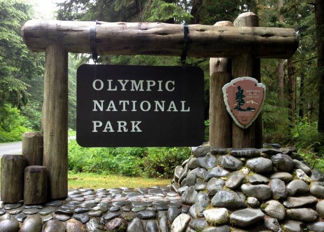 A carved park entrance sign hangs from a log frame atop a base made from river rocks.