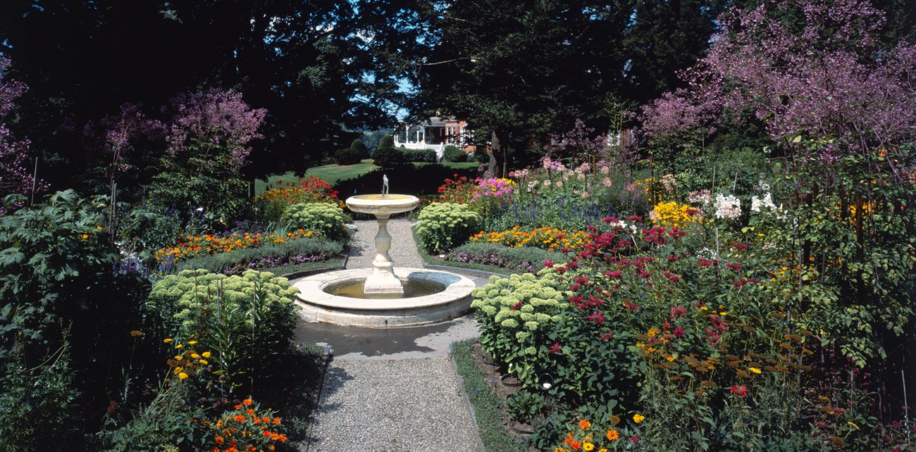 2500-MABI_formal_garden A fountain stands at the axis of a gravel footpath in a formal garden, dense with colorful flowers and shrubs.