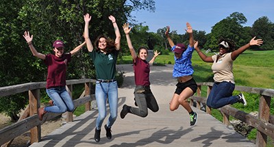 Five young women smile and reach into the air as they jump above a wooden footbridge