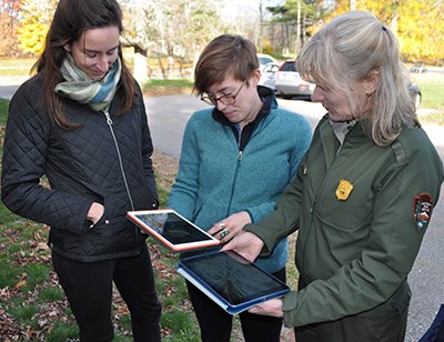 400-Plant Records Management 1 Three women, one in NPS uniform, look at two digital tablets in an autumn landscape.