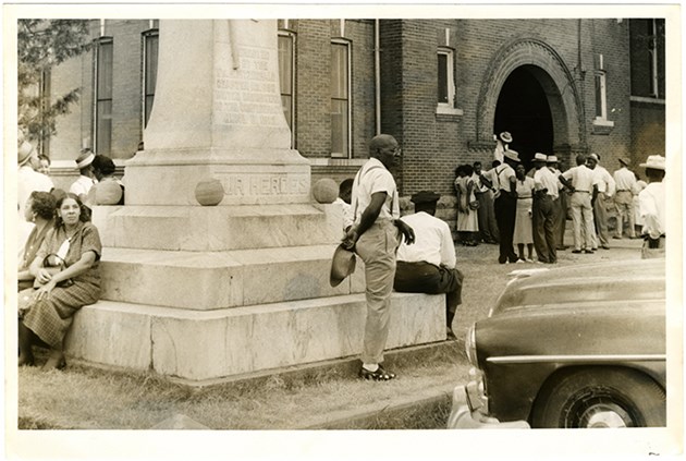 Men and women wait outside; some lean on a monument others gather around a building.