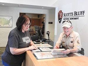 A man in a volunteer uniform shows a visitor a map at a visitor center desk.