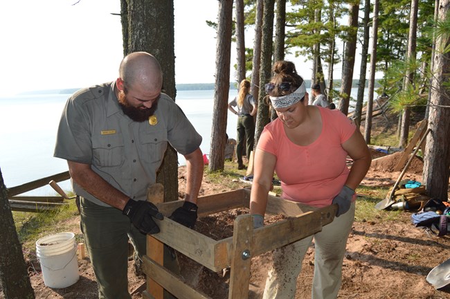 Two people stand among evergreen trees sifting dirt through a shaker screen. Other people and a lake are visible in the background.