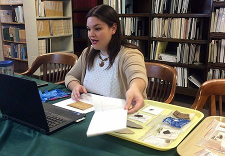 Woman seated in front of a laptop computer holding a piece of paper with a tray of artifacts to her left on a table. Shelves of books are behind her.