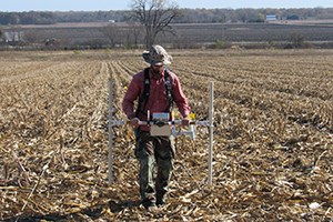 Archeologist uses a handheld magnetic gradiometer to survey a harvested corn field.