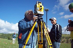 An archeologist sets up a robotic total station on a tripod in a grassy field while four other crew members hold equipment in the background. Green, rolling hills are in the distance.