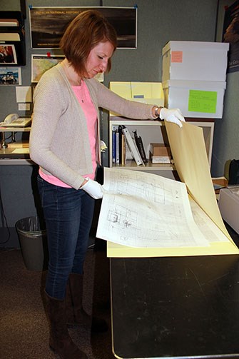 Standing woman opens a large manila folder and places an oversized map inside. She is wearing white gloves.