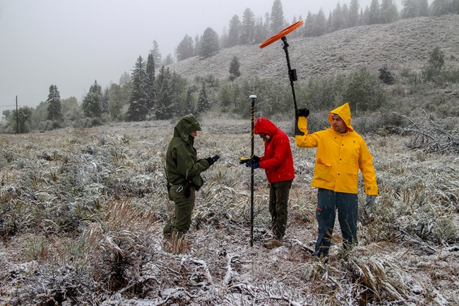 3 archaeologists working on a snowy day; the left archeologist takes notes while the archeologist at center uses a GPS unit mounted on a pole, while the other holds a magnetic susceptibility meter in the air.