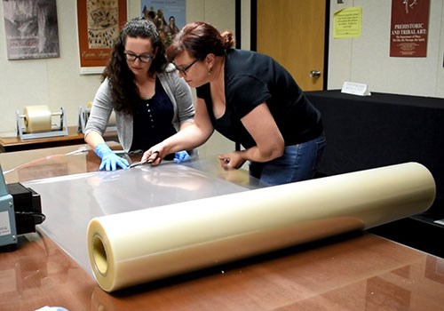 Two women hold and cut a large roll of plastic sheeting to size on a large wooden table