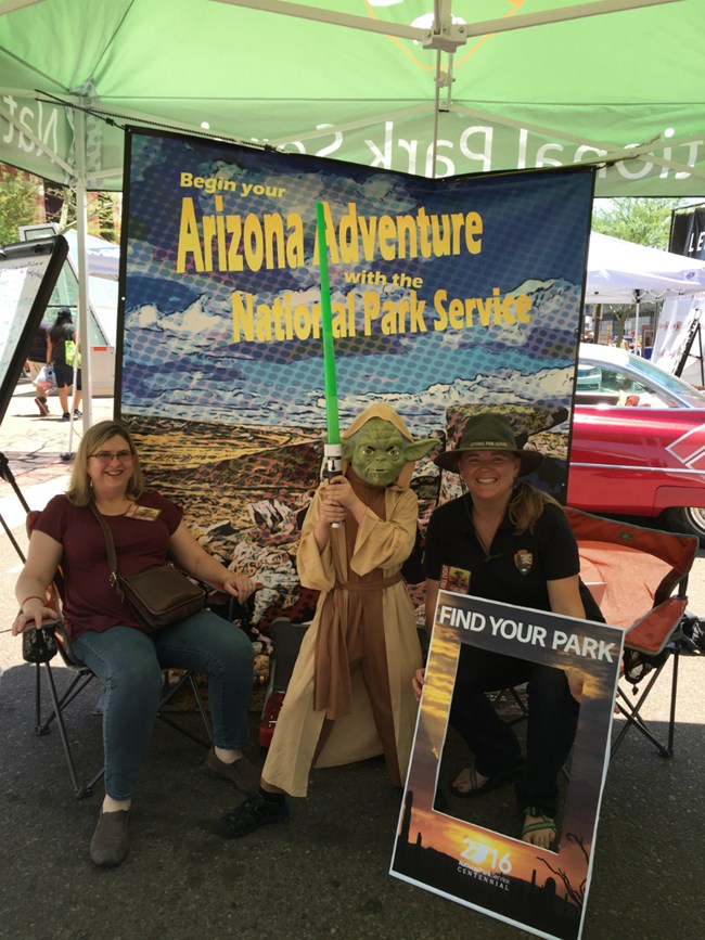 An NPS ranger with a kid dressed as Yoda in front of a pixelated backdrop picture of Petrified Forest.