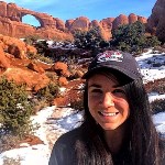 A smiling woman with long hair and ballcap in front of snow covered red rocks and arch.