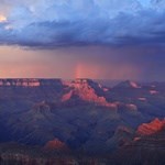 Dark clouds hover over a deep canyon turned red by sunset.