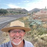 A smiling man in uniform flat hat and glasses in front of a road and entrance sign to Tonto National Monument