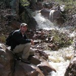 A man in ball-cap sits on a rock by a flowing creek.