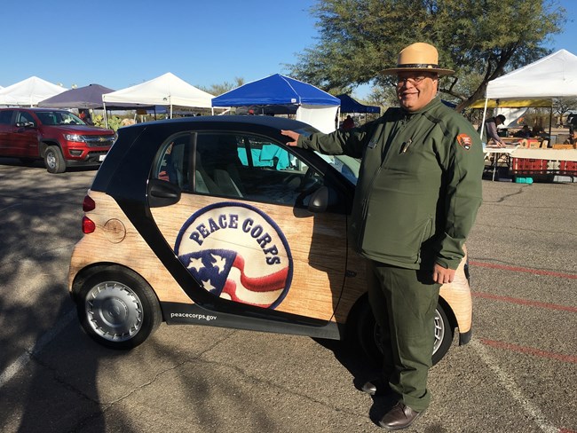 Karl Pierce with Peace Car_11192017 A man in green uniform and hat stands in front of a small electric car.