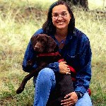 A smiling woman with long hair and glasses holding a brown dog with a red collar.