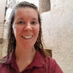 A woman in red wine colored polo shirt smiles while standing in front of an old looking adobe buidling.