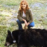 A blonde woman squats behind a black bear; the bear is laying on the ground, asleep, a radio collar around it's neck.