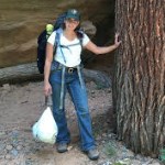 Rodriguez in the field A woman in green ball cap and hiking backpack stands with hand on a tree.