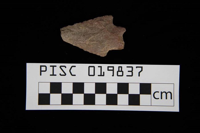 stone projectile point with checkered centimeter scale below on black background.