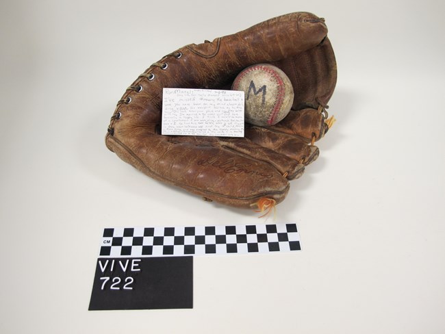 leather baseball glove and white baseball with a handwritten explanation card above a checkered centimeter scale.