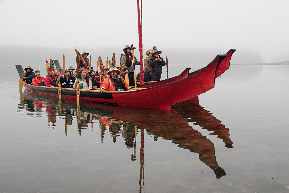 A group of people, dressed in full Tlingit regalia, arrive in a canoe. There is a reflection on the water.