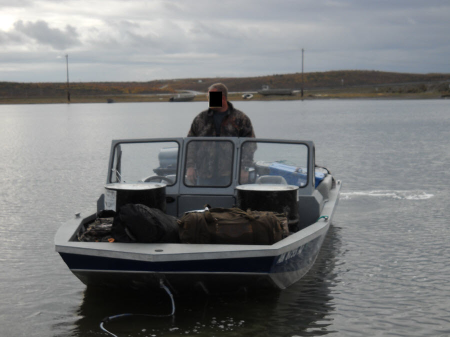 Figure 1 - Jet boat used by Silvis to transport unlawfully taken game, hunters and gear into and out of the Preserve. A jet boat sits in the water . A man, whose face is blacked out stands in the boat. There are gear bags visible in the front of the boat.