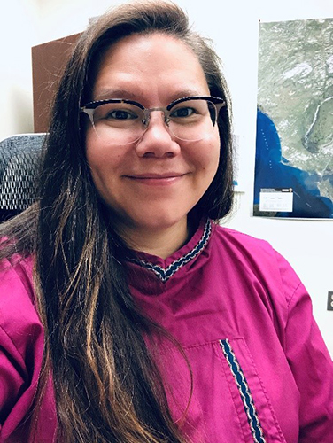 A profile picture of a woman smiling. She has long dark hair and wears glasses. She is wearing a bright shirt with trim.