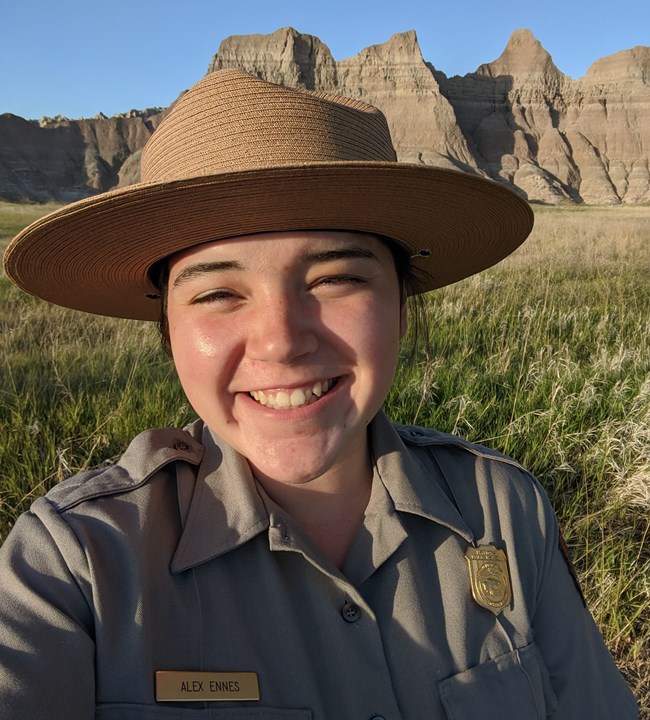 a female park ranger wearing a tan flat hat, grey button-down, and badge smiles in front of rugged, buff badlands buttes.