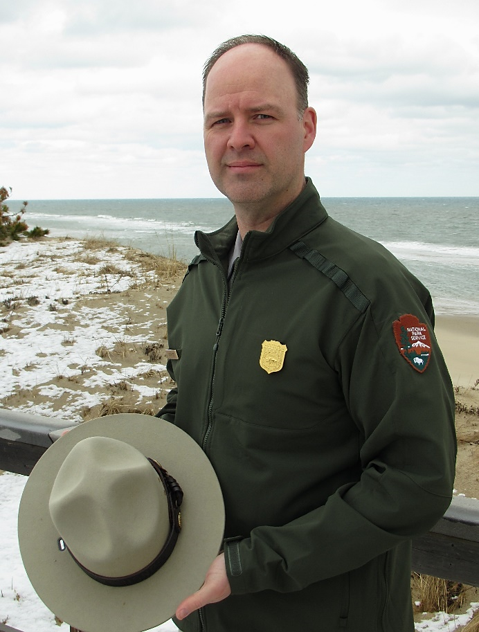 Jason Taylor has been selected as Superintendent for Klondike Gold Rush National Historical Park, in Skagway, Alaska. Jason Taylor stands in uniform facing the camera. He is holding his hat in his hands and the ocean is seen in the background.