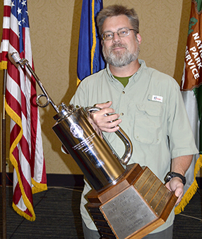 Arkansas Park Group wins annual Midwest Region Fuels Award Fenn Wimberly holds a driptorch trophy in front of flags.