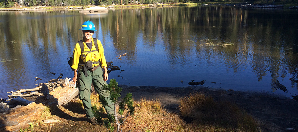 Jen Gibson wears personal protective equipment standing on a lakeshore with a coniferous forest in the background.
