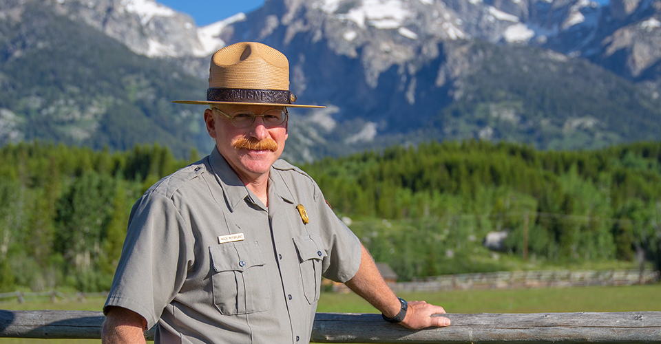 Mack McFarland stands against a fence with the Grand Teton mountain range as a backdrop.