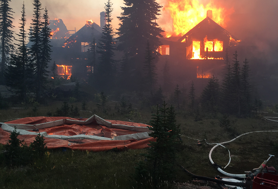 A structure burns in a forest with a portable water tank, hose, and pump in the foreground.