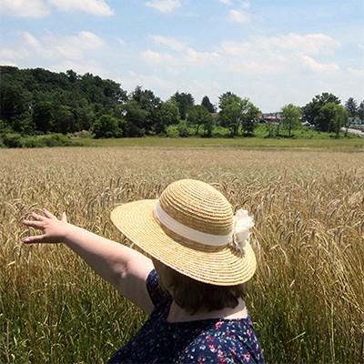 Woman in sun hat gestures over wheat field, trees and road in distance