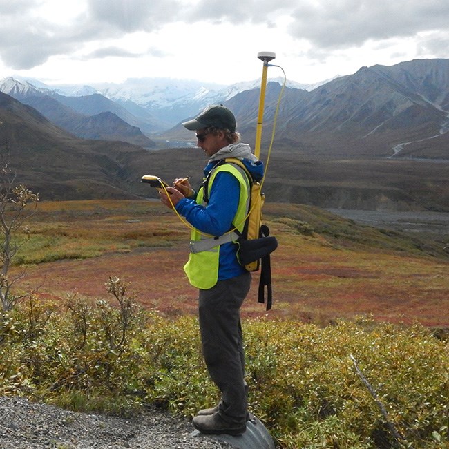 Man with large backpack with column rising over his head takes notes on rugged mountain landscape