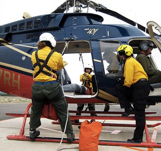 A crew member leans out of a helicopter with a rope while standing on the landing skid.