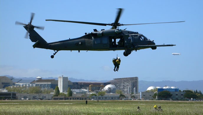 Two people hang from a hoist line below a large hovering helicopter.
