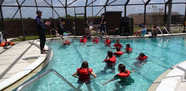 Water ditching and survival A group of students stands in a pool with life vests listening to a person talk from the edge of the pool.
