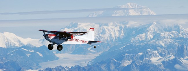 lt= Fixed wing single engine aircraft flying under blue skies and above snowy mountains.