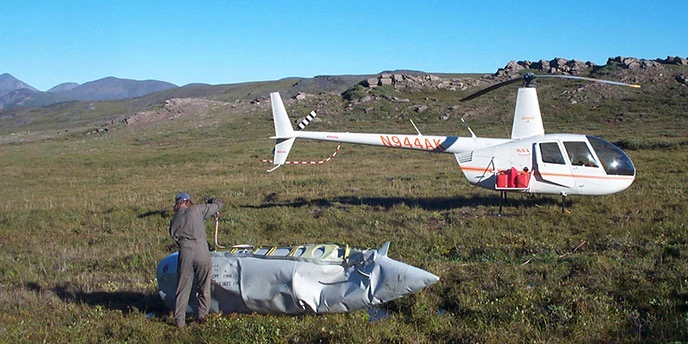 A person in a flight suit stands near a fuel container while a helicopter is waiting nearby.