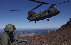 Partners-army-aviation-support-mount-rainier A person in a green flight suit hunches on the ground while a helicopter takes off nearby in a rocky mountainous area.