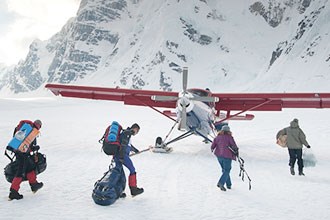 lt= A group carrying packs walks toward an airplane on a glacier with snowy mountains in the background.