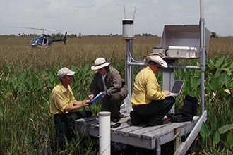 lt= Scientists on a platform conduct studies in a swampy area while a helicopter waits nearby.