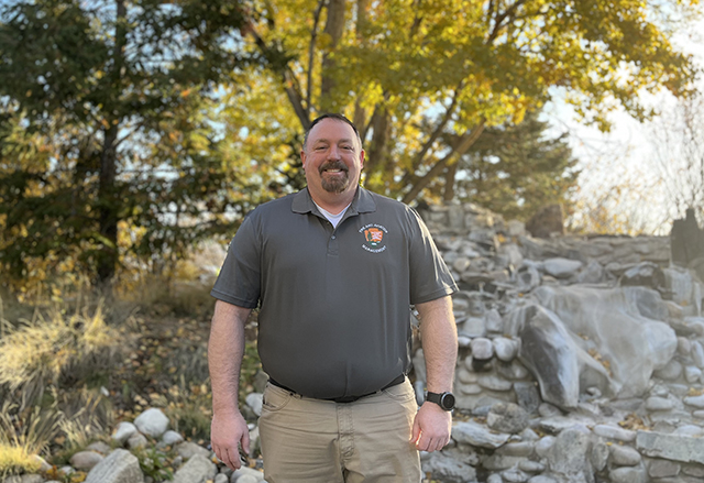 Brian Sutton Man stands smiling in front of trees and rocks.