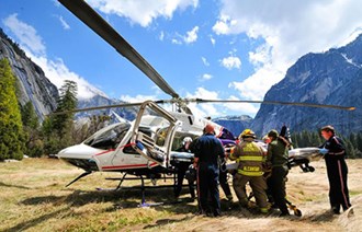 lt= A group loads a stretcher into a helicopter in a meadow surrounded by mountains.