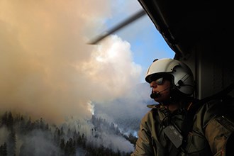 lt= A woman in a flight helmet looks out the door of a helicopter on mountain filled with smoke.