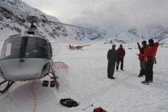 lt= group stands outside of helicopter on snowy field with an airplane in the background