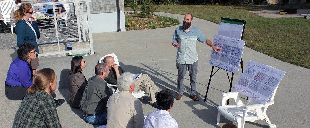 A group of people sit on concrete steps outside watching a person present while pointing at an easel A group of people sit on concrete steps outside watching a person present while pointing at an easel
