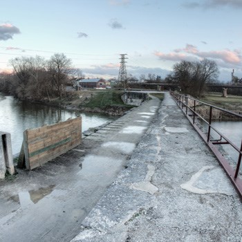 An abandoned bridge crosses over a river under a cloudy grey sky.